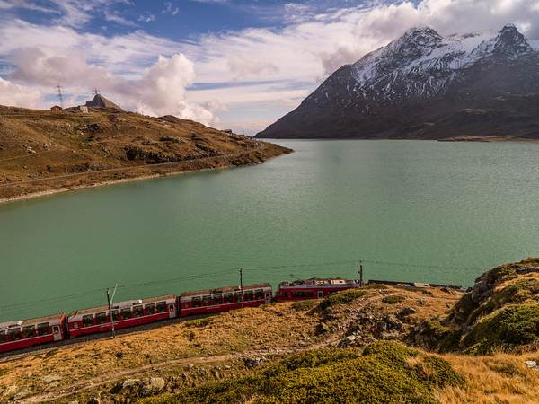 Der Bernina Express der Rhätischen Bahn unterwegs entlang des Lago Bianco auf dem Berninapass. Im Hintergrund der Sassal Mason.