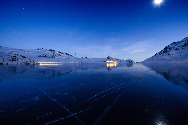 Abendstimmung über dem zugefrorenen Lago Bianco auf dem Berninapass mit der Rhätischen Bahn im Hintergrund