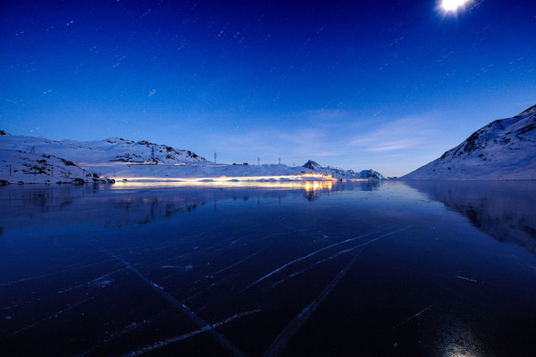 Abendstimmung über dem zugefrorenen Lago Bianco auf dem Berninapass mit der Rhätischen Bahn im Hintergrund